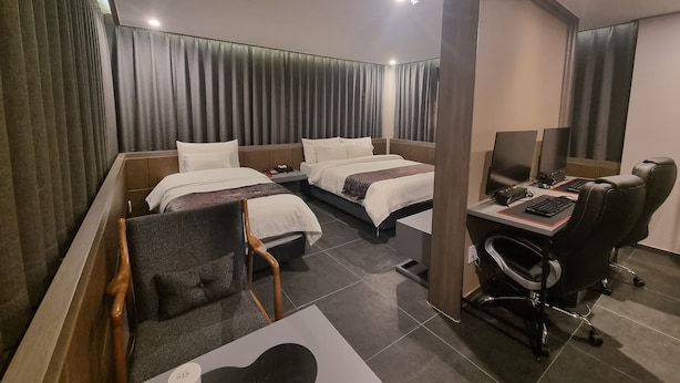 A hotel room with two beds, a desk with a computer, a chair, and a TV. - Yeow-B (Jinju)