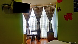 A hotel room with a bed, a TV mounted on the wall, a chair, a small table, and a window with curtains. - C & L Suites Inn (Dumaguete)