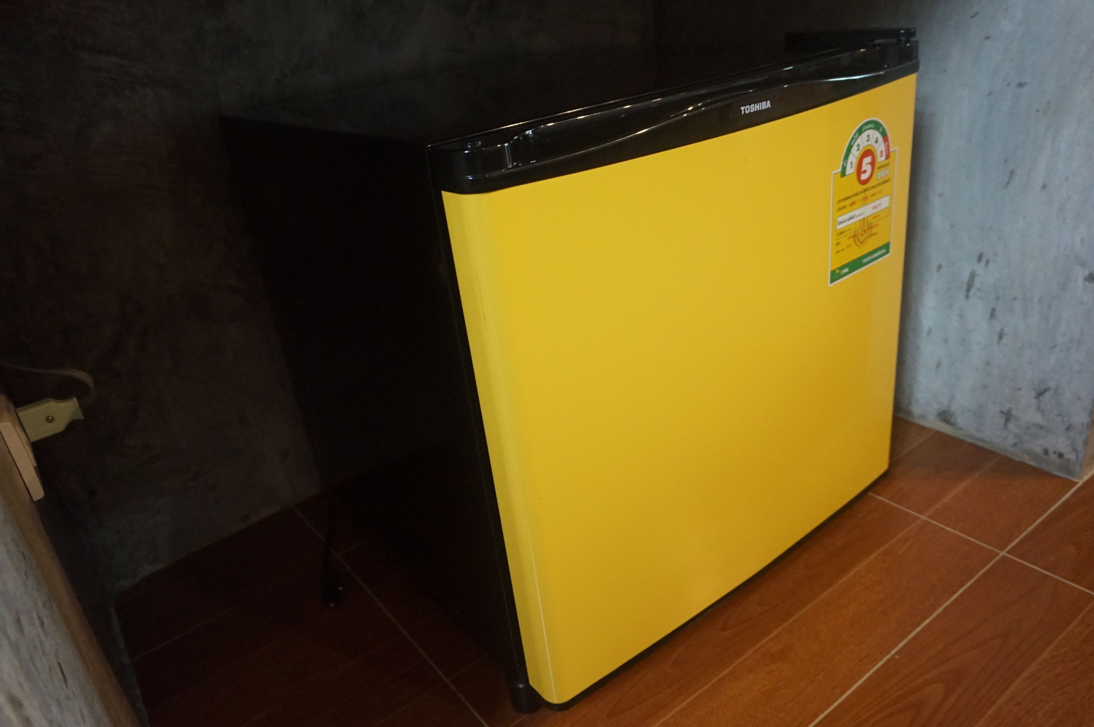 Un electrodoméstico amarillo pequeño con una pegatina, ubicado en una habitación con suelo de baldosas y pared de hormigón.