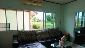 A living room with a black leather sofa, a glass coffee table, and a view of a garden through large windows. - A Nice Resort & Villa (Buriram)