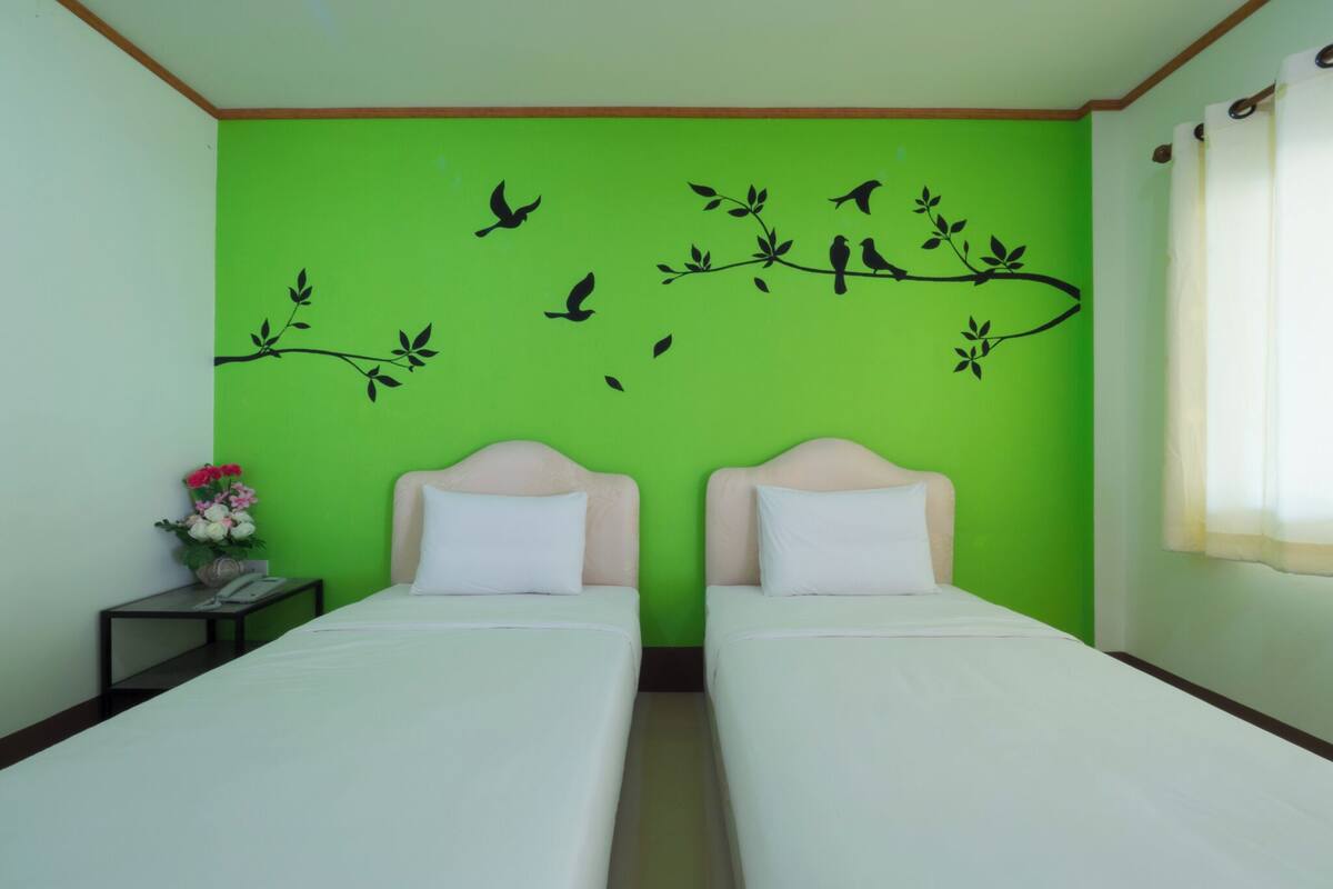 Une chambre avec deux lits, un mur vert orné d’une fresque représentant un oiseau et une branche, une petite table avec un vase de fleurs et une fenêtre avec des rideaux.