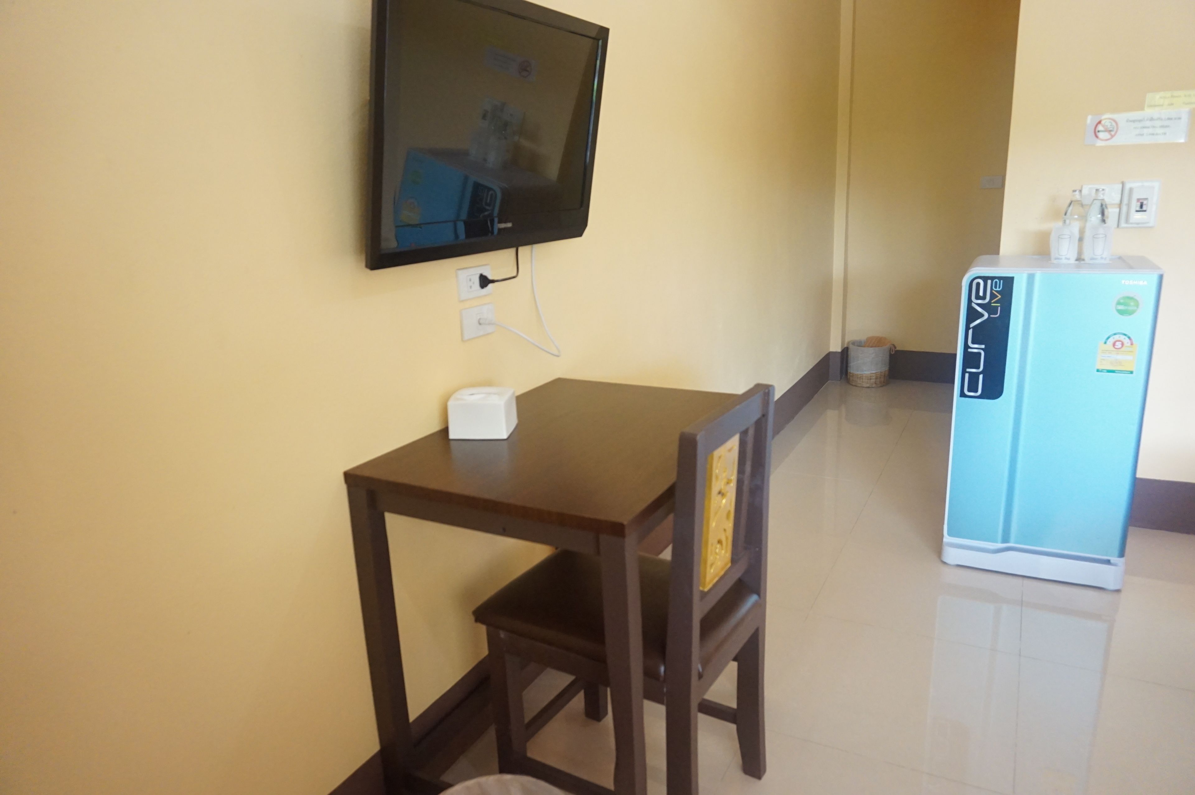 A small room with a wooden table and chair, a television mounted on the wall, and a blue refrigerator with a water dispenser.
