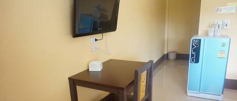 A small room with a wooden table and chair, a television mounted on the wall, and a blue refrigerator with a water dispenser.