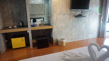 Una habitación de hotel con cama, televisor montado en la pared, una mesita con hervidor, tazas y botellas, un minibar amarillo, un reposapiés negro y una cesta de mimbre.