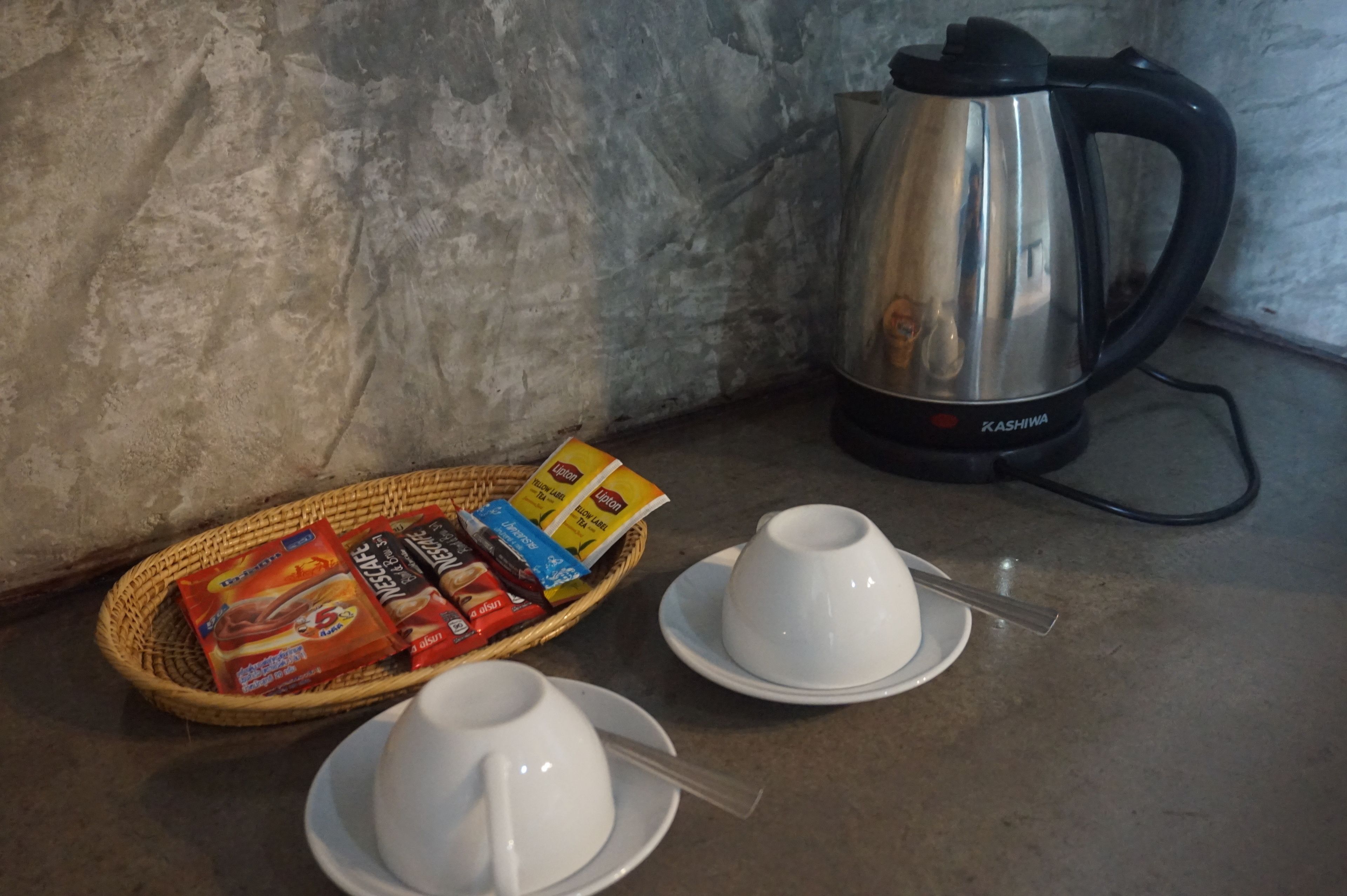 A kitchen counter with a stainless steel electric kettle, a wicker basket with tea bags and a packet of instant coffee, and two white cups with saucers.