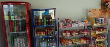 Una máquina expendedora con diversas bebidas y aperitivos, un refrigerador de Coca-Cola y un estante lleno de productos envasados.