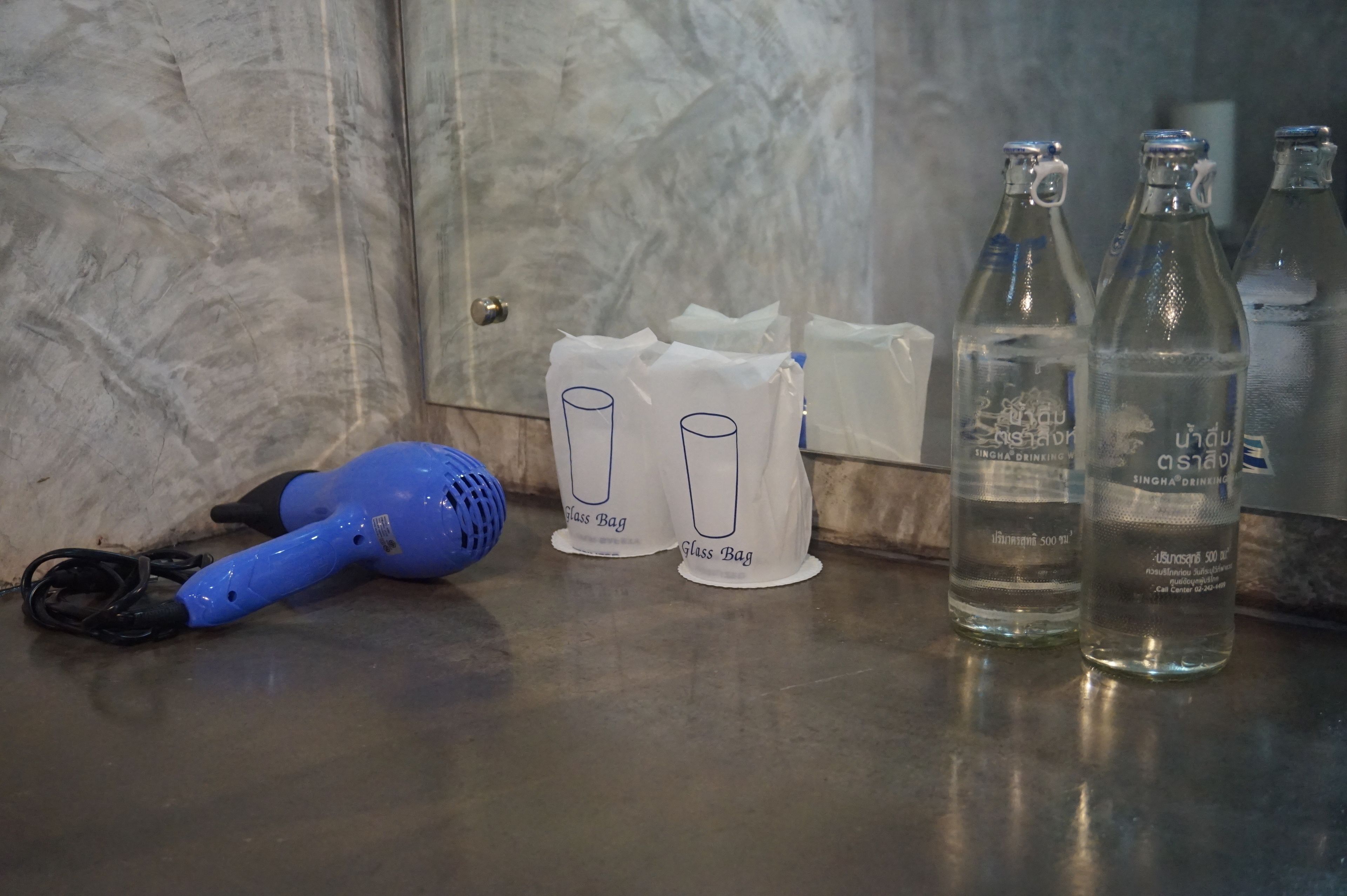 Une salle de bain avec un mur en marbre, un sèche-cheveux bleu, trois sacs en verre et deux bouteilles d’eau.