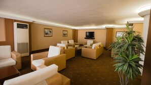 Et hotelværelse med beige vægge, et fladskærms-tv monteret på væggen, flere siddeområder med beige sofaer og lænestole, en stor stueplante og et tæppe på gulvet.