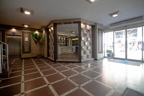 Vestíbulo de hotel con suelo a cuadros, un ascensor, una recepción y un gran ventanal que da al exterior.