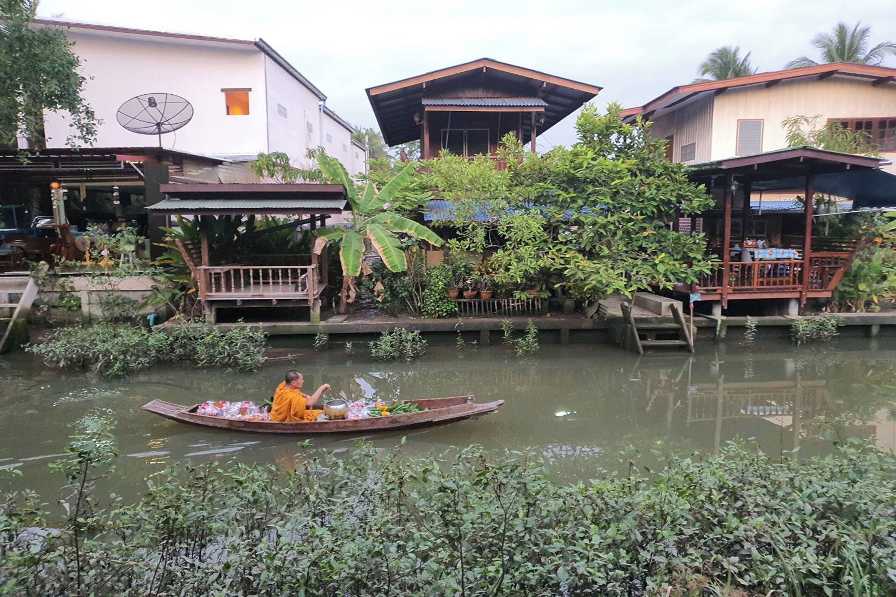 A river with a boat carrying a monk and offerings, surrounded by buildings and lush greenery.