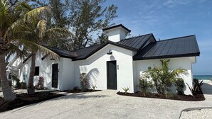 A modern white house with a black roof, surrounded by palm trees and a clear view of the ocean.