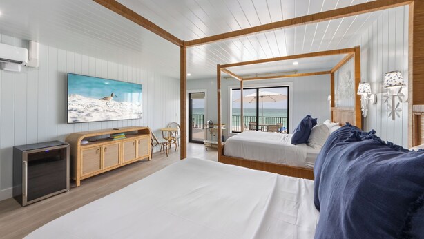 A modern bedroom with a large bed, a TV mounted on the wall, a wooden headboard, and a view of the sea through the balcony.