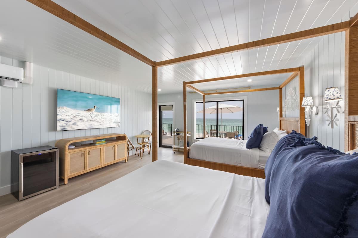 A modern bedroom with a large bed, a TV mounted on the wall, a wooden headboard, and a view of the sea through the balcony.