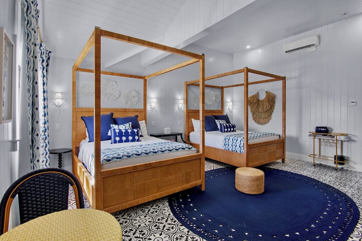 A four-poster bed with a wooden frame, a round woven stool, and a patterned rug.