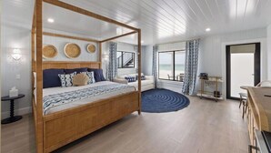 A spacious bedroom with a four-poster bed, a sitting area with a sofa, a dining table, and a view of the sea through large windows.