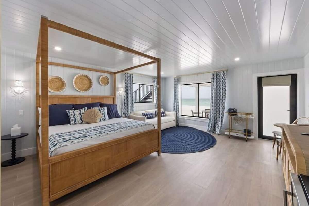 A spacious bedroom with a four-poster bed, a sitting area with a sofa, a dining table, and a view of the sea through large windows.