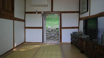 Una habitación japonesa tradicional con suelo de tatami, una puerta corrediza que da a un jardín, un armario de madera y un televisor a la derecha.
