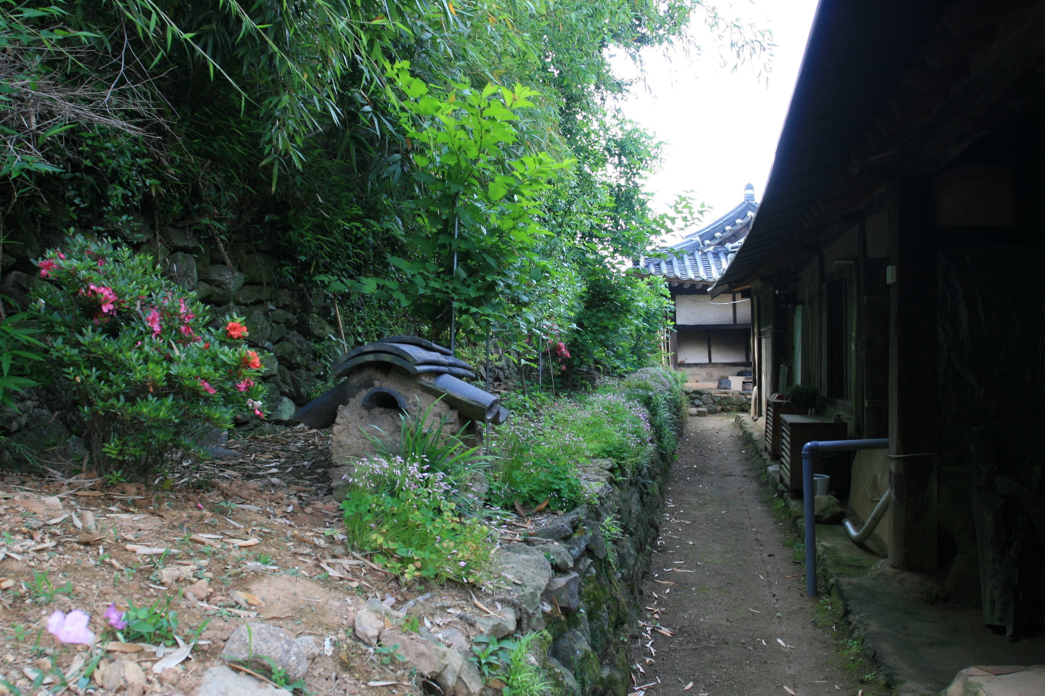 A traditional Japanese garden with a stone bridge, lush greenery, and a pathway leading to a building with a tiled roof.