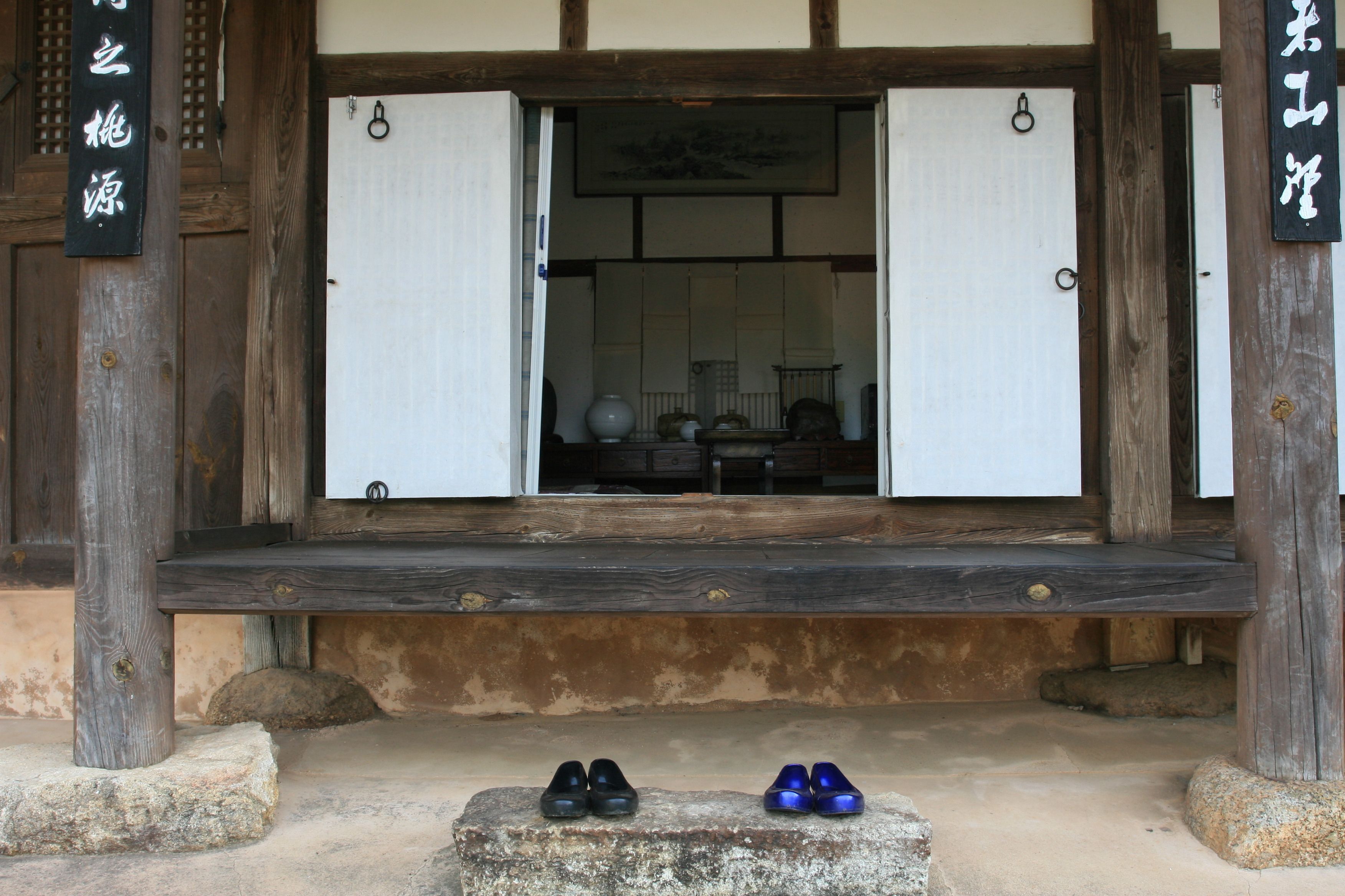 A traditional Japanese building entrance with a wooden structure, a stone platform, and two pairs of shoes on the ground.
