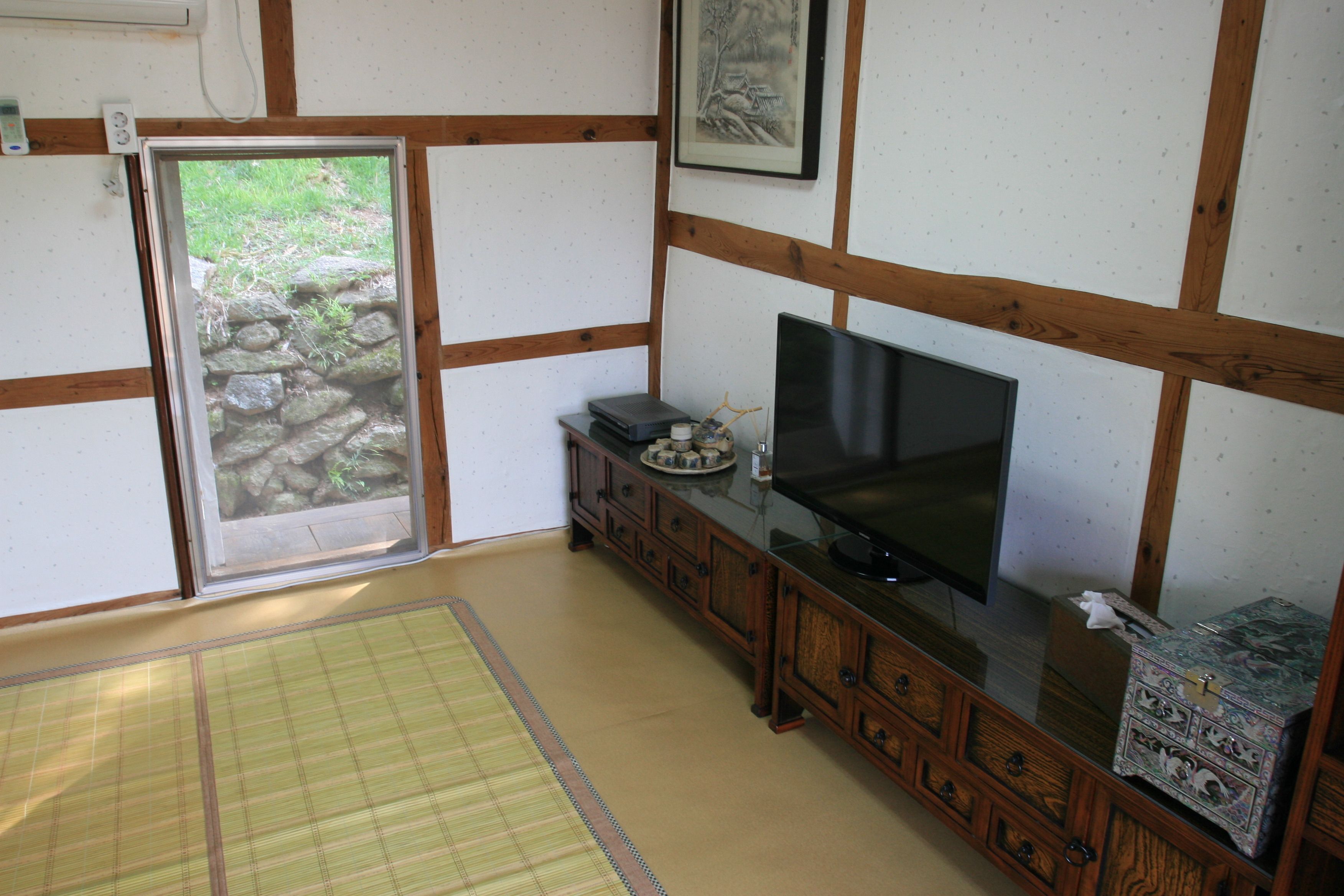 A traditional Japanese room with tatami flooring, a sliding door leading to a garden, a television on a wooden cabinet, and a framed artwork on the wall.