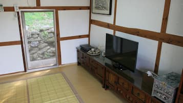 A traditional Japanese room with tatami flooring, a sliding door leading to a garden, a television on a wooden cabinet, and a framed artwork on the wall.