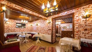 A room with brick walls, wooden ceiling beams, and a red carpet. There are tables and benches, and a chandelier hangs from the ceiling. - Akcasu Konuk Evi (Safranbolu)