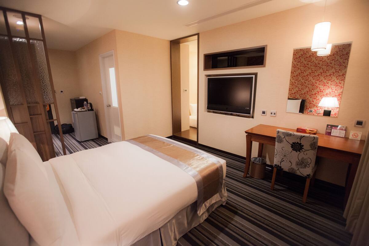 A hotel room with a bed, a desk, a chair, a TV, a mirror, and a small kitchenette.