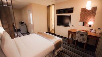 A hotel room with a bed, a desk, a chair, a TV, a mirror, and a small kitchenette.
