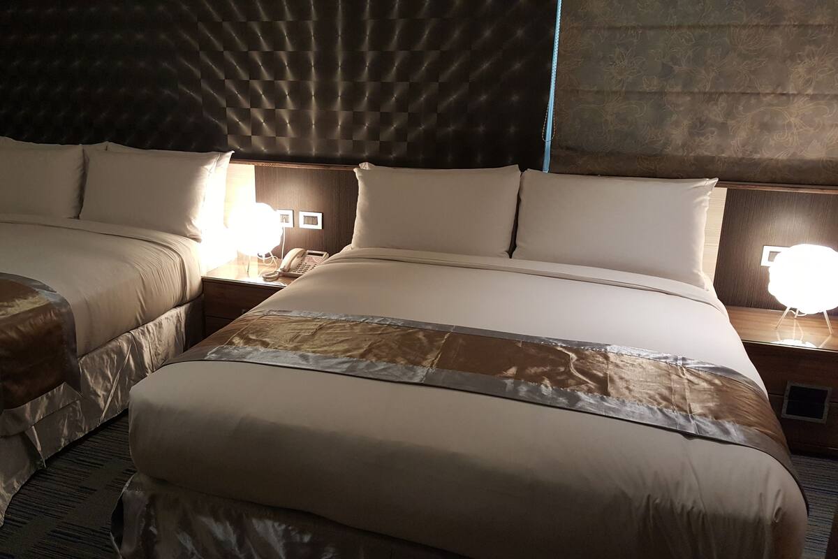 A hotel room with two beds, each with a headboard, bedside tables with lamps, and a large window with curtains.