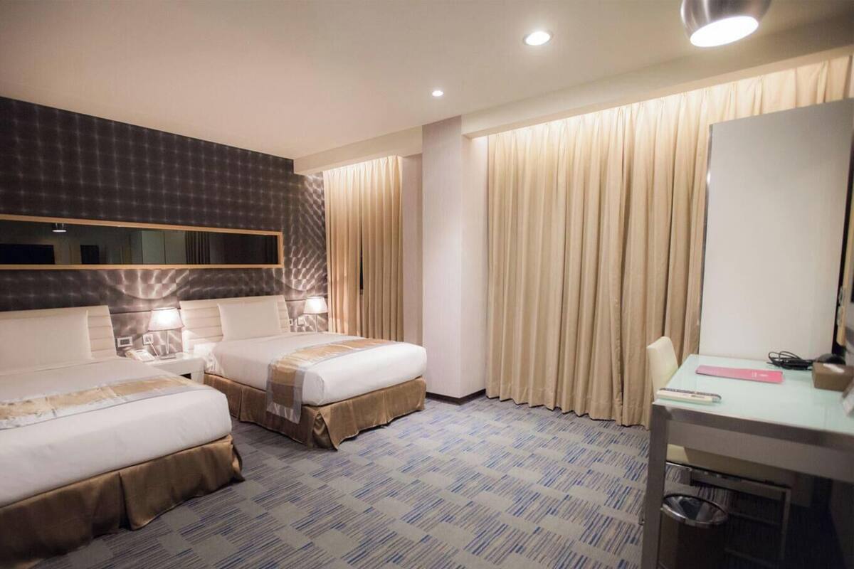 A hotel room with two beds, a desk, a mirror, and a window with curtains.