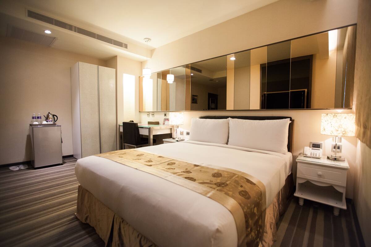 A hotel room with a large bed, a desk, a chair, a wardrobe, and a small desk with a lamp.