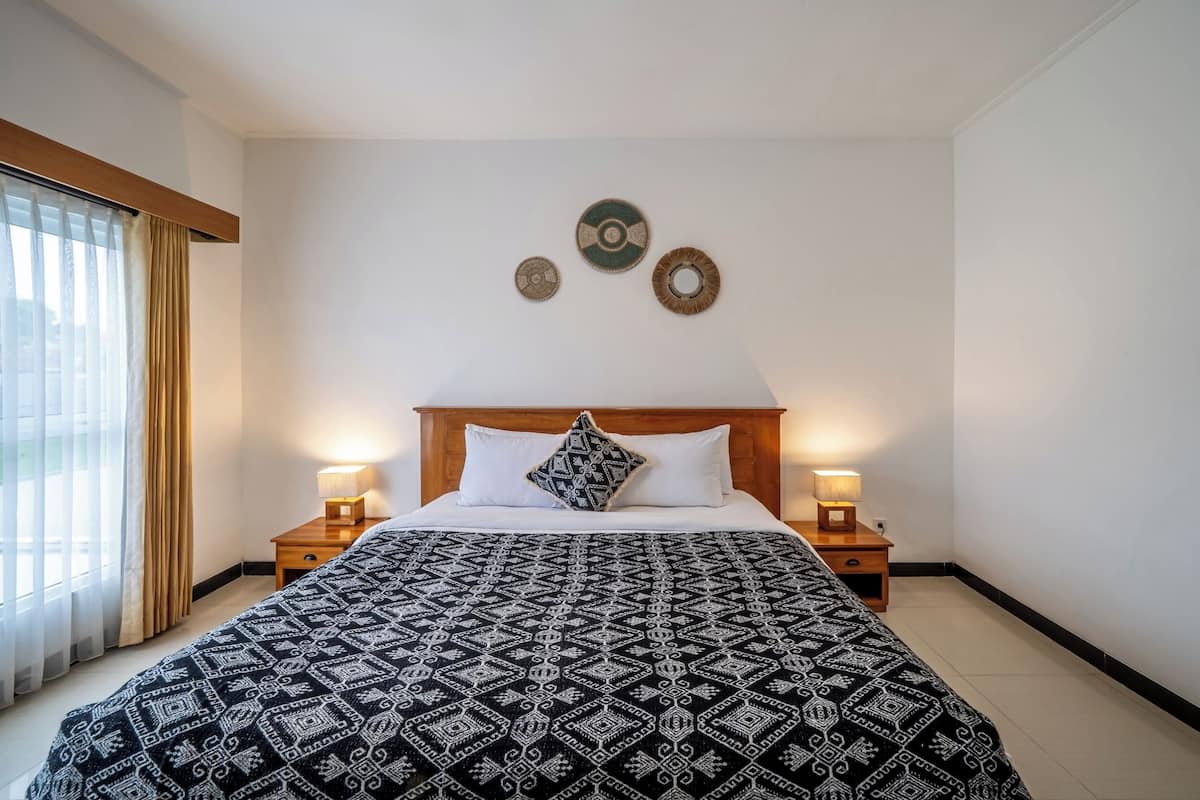Una habitación de hotel con una cama grande, dos mesitas de noche con lámparas, un ventanal con cortinas y adornos en la pared.