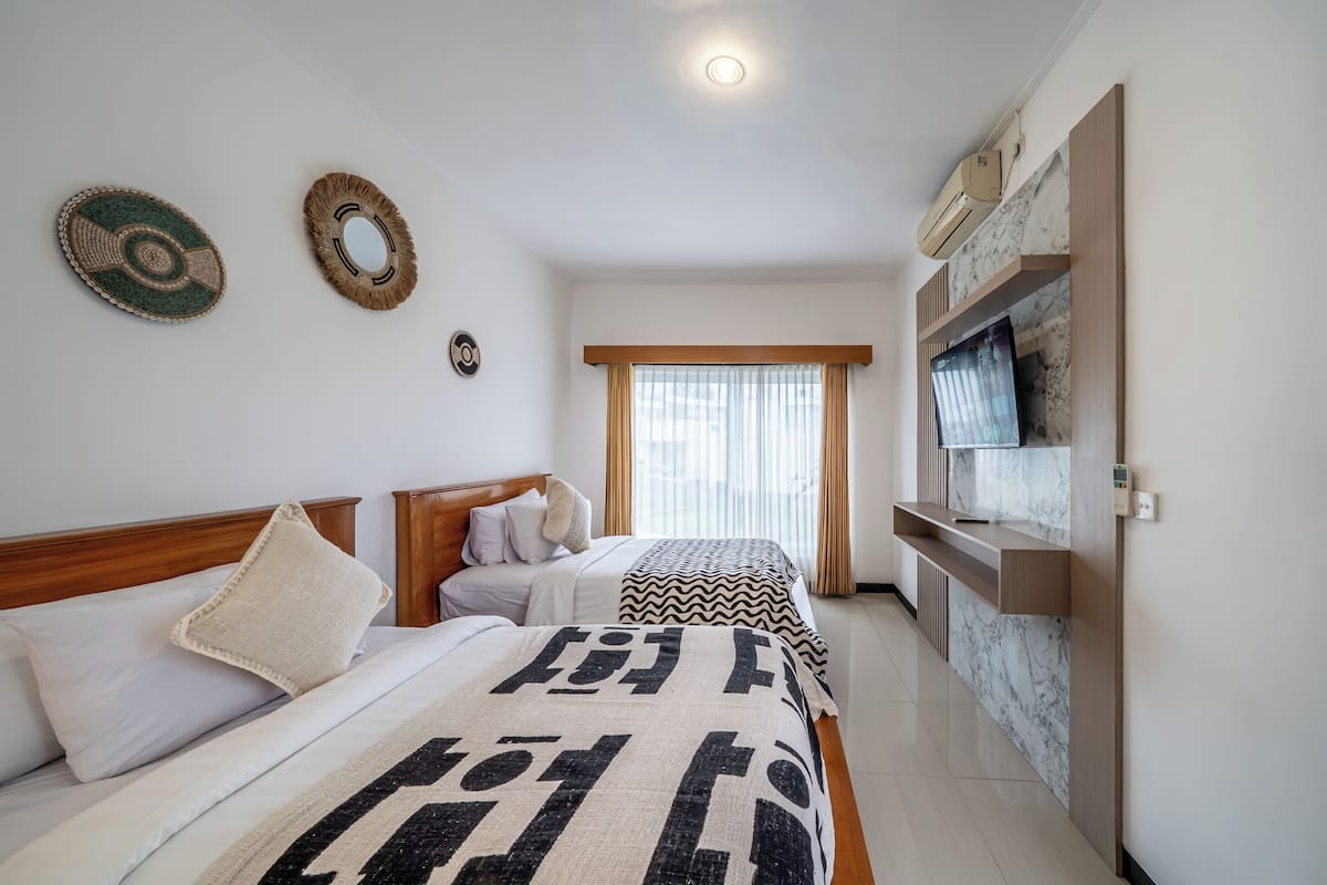 Habitación de hotel con dos camas, cabecera de madera, televisor montado en la pared y ventana con cortinas.