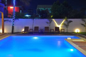 A nighttime swimming pool with blue lighting, surrounded by lounge chairs and a white wall with small lights. - Paragon Hotel (Mandalay)