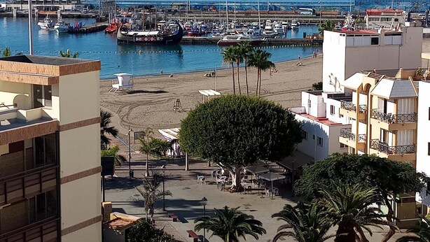 Una marina costera con barcos amarrados, una playa de arena y edificios con balcones que dan al mar.
