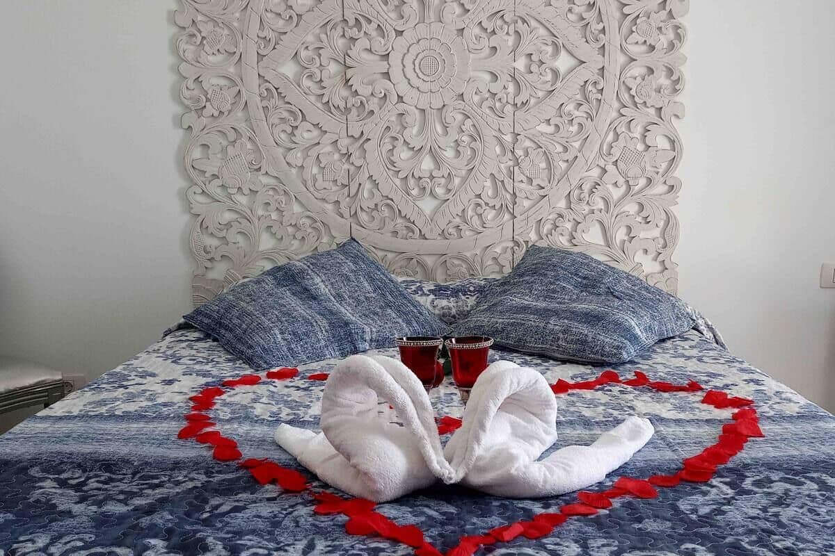 Un lit agrémenté d’une décoration en forme de cœur, composée de serviettes et de pétales de roses rouges, ainsi que de deux tasses rouges et d’une tête de lit décorative.