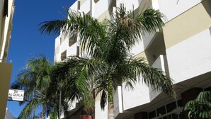 Un edificio de varios pisos con fachada blanca y balcones. Palmeras se ven al frente.