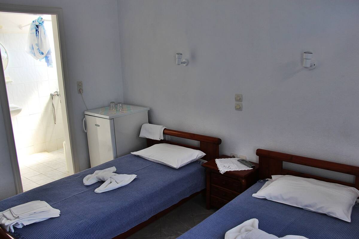 Une chambre d’hôtel avec deux lits simples, une petite table de chevet avec une lampe, une salle de bain avec douche et un lavabo.