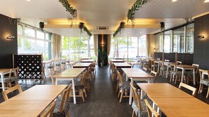 Un restaurante espacioso con largas mesas y sillas de madera, amplias ventanas con luz natural y plantas colgando del techo.