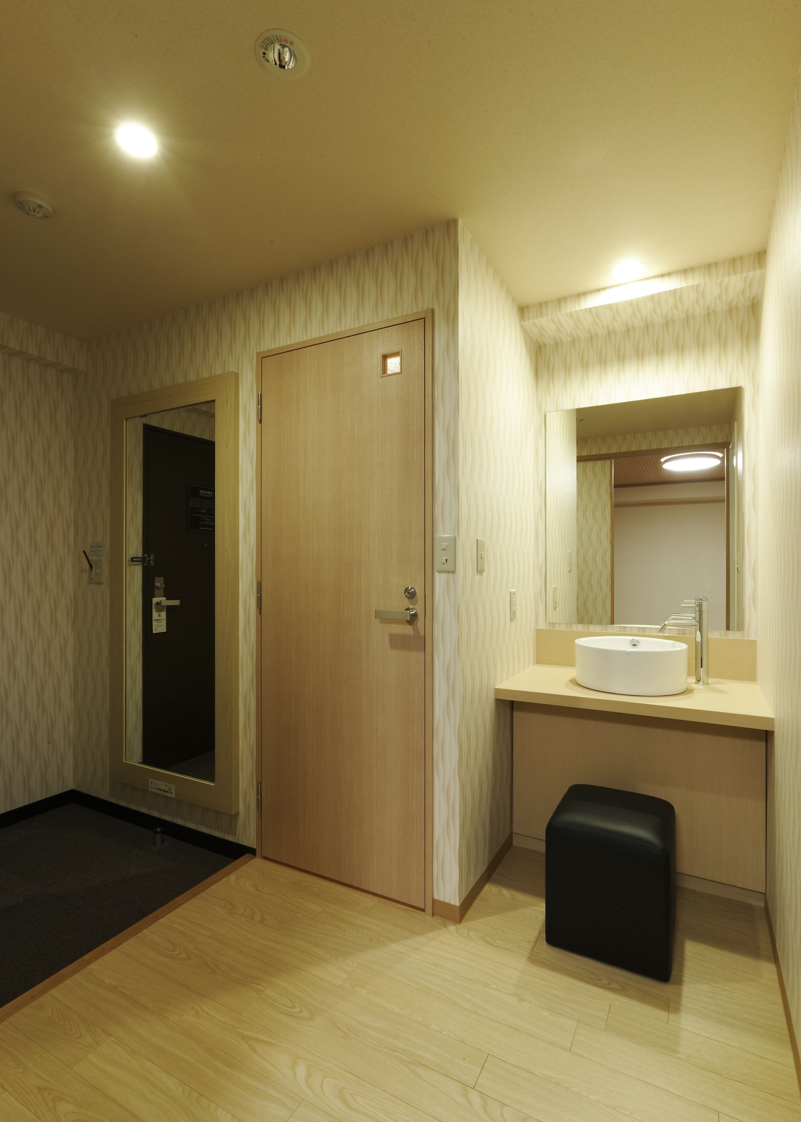 A hotel room with a wooden door, a small mirror above the sink, a black stool, and a light-colored floor.