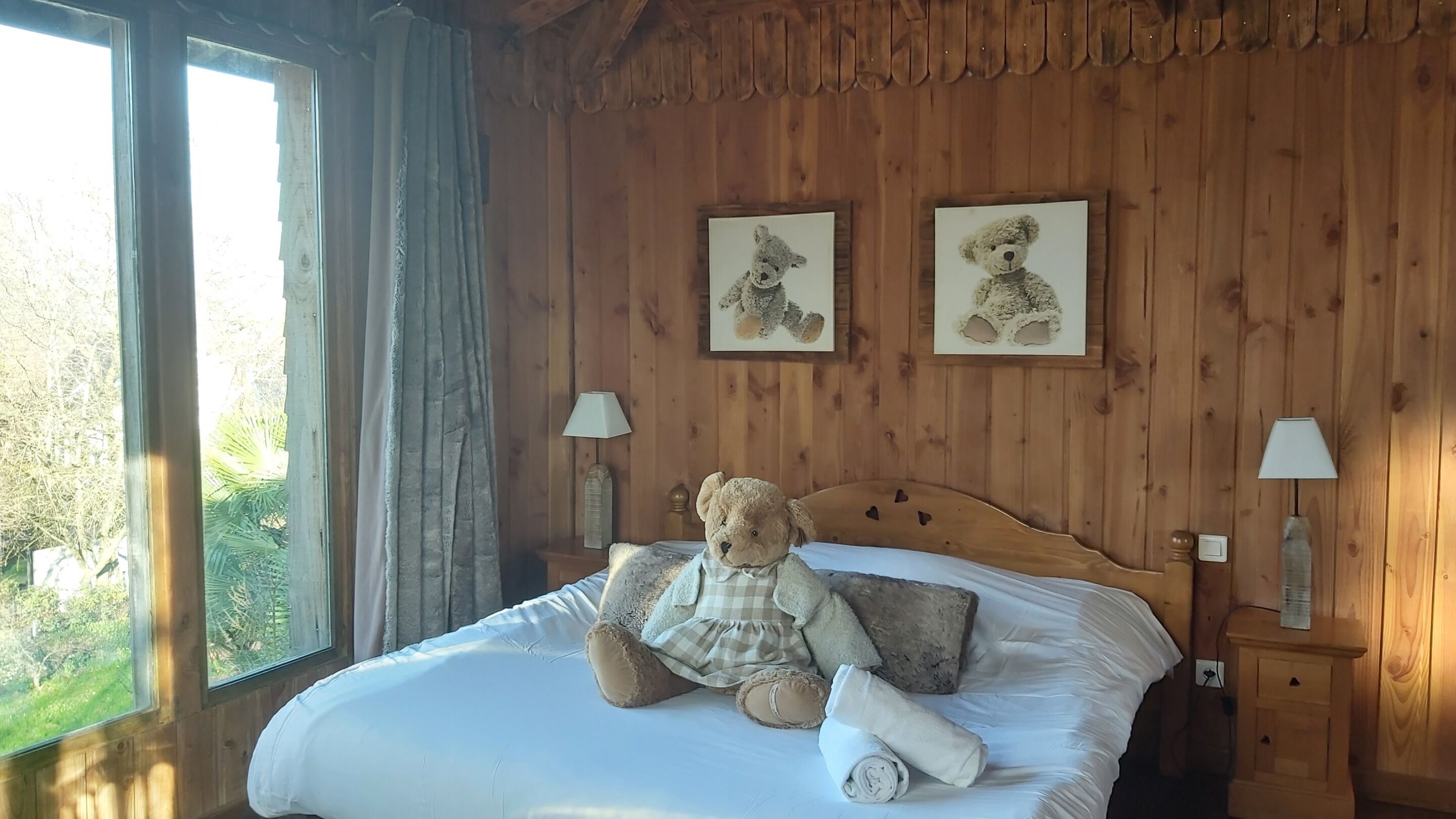 Ein Zimmer mit Holzboden, einem Bett, zwei gerahmten Bildern, einem Teddybären und einer Lampe.