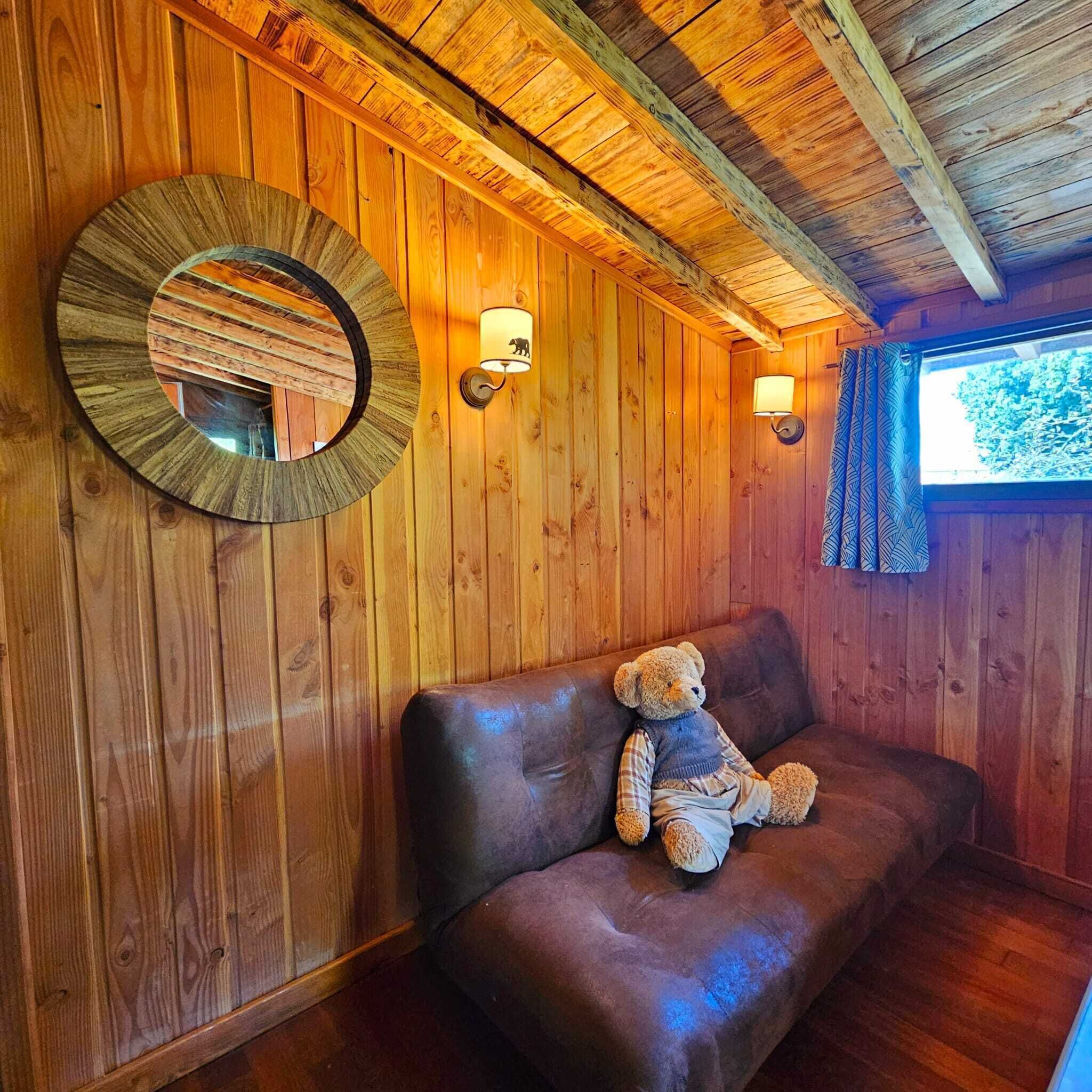 Interior de una cabaña de madera con un sofá de cuero marrón, un osito de peluche, un espejo redondo y dos lámparas fijadas a la pared.