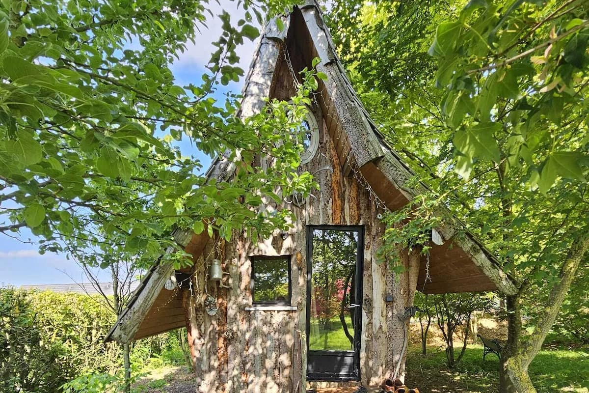 Een rustieke boomhut met een houten deur, een klein terras en een metalen tafel met stoel.