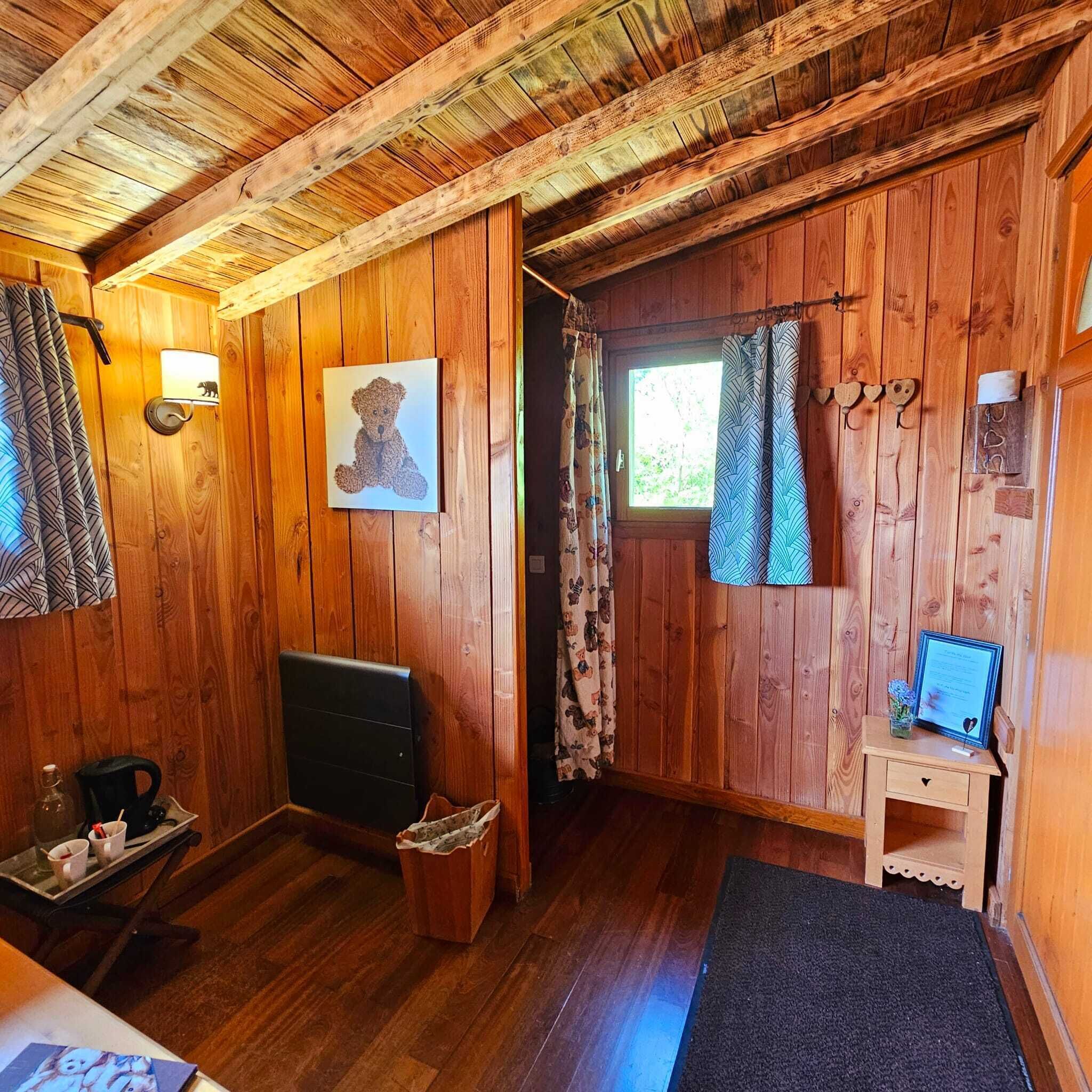 En stuga med träpanel på insidan, inredd med en säng, ett skrivbord, en stol, ett fönster med gardiner och en väggdekoration med björntema.