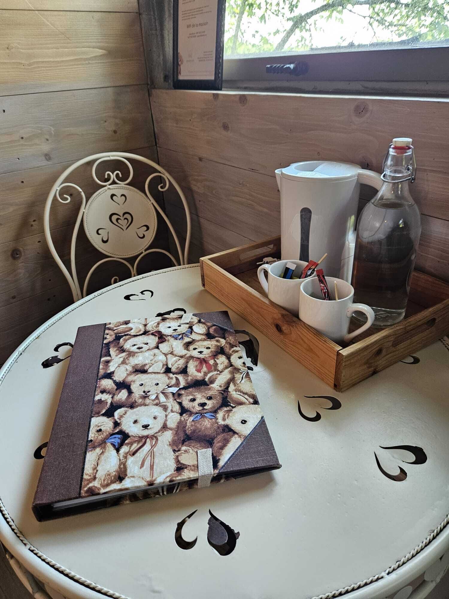 A wooden interior with a round table, a chair with heart designs, a tray with a mug, a bottle, and a book with teddy bear illustrations.