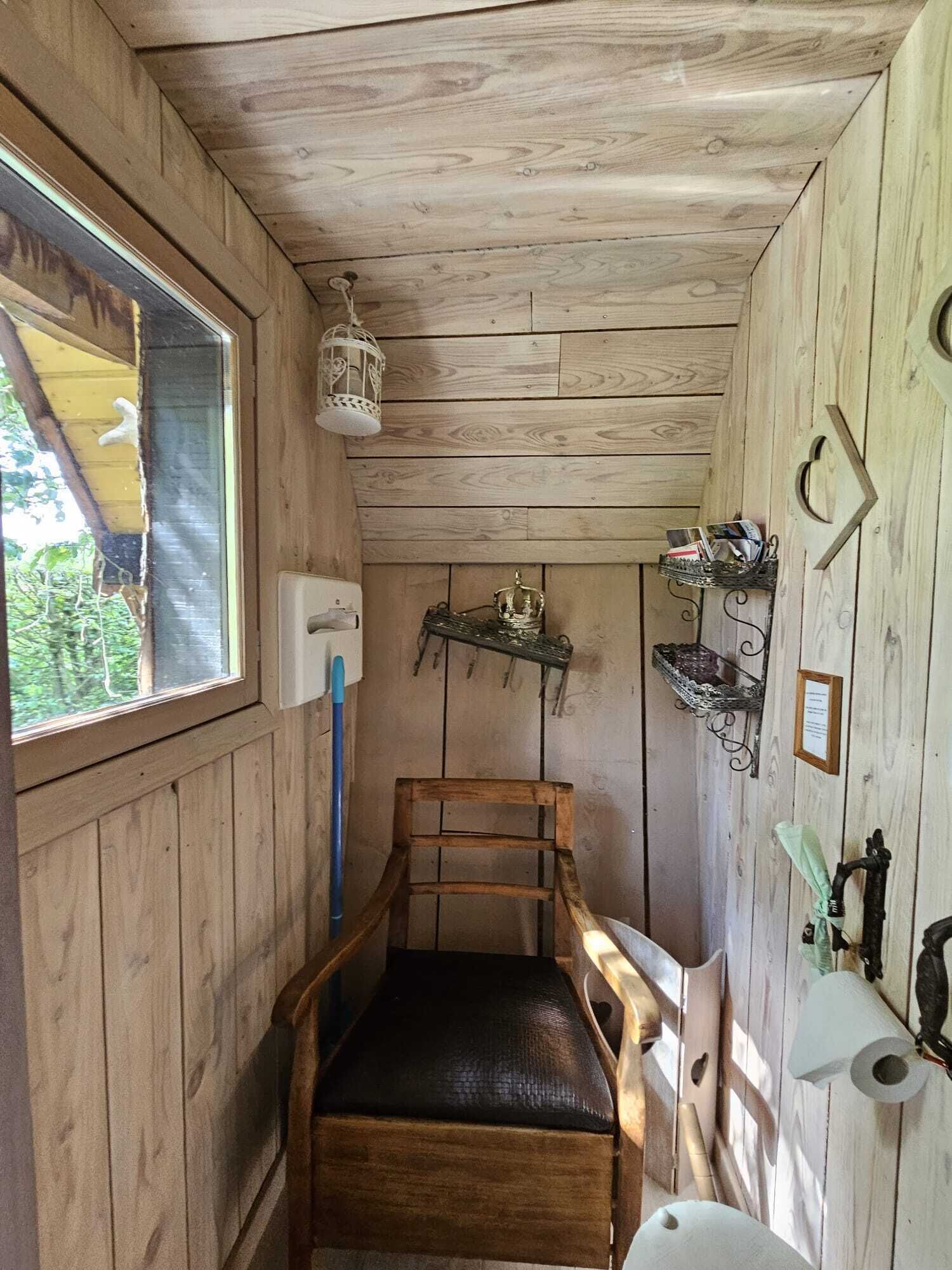 A wooden cabin interior with a wooden chair, a small shelf with books, a lantern, and a window with a view of greenery.