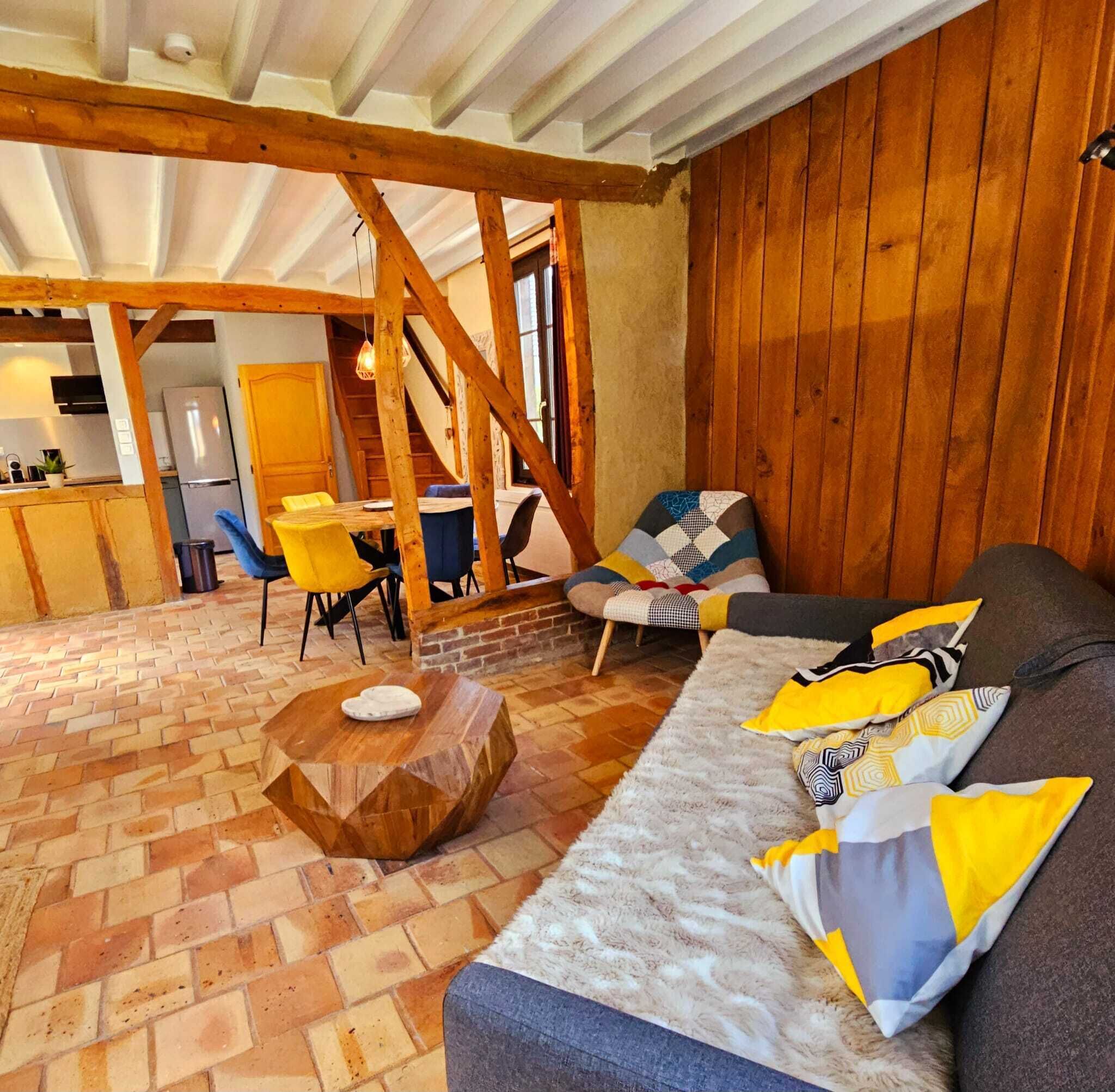 A cozy living room with a brick floor, wooden beams, a sofa with patterned cushions, and a dining area with chairs.