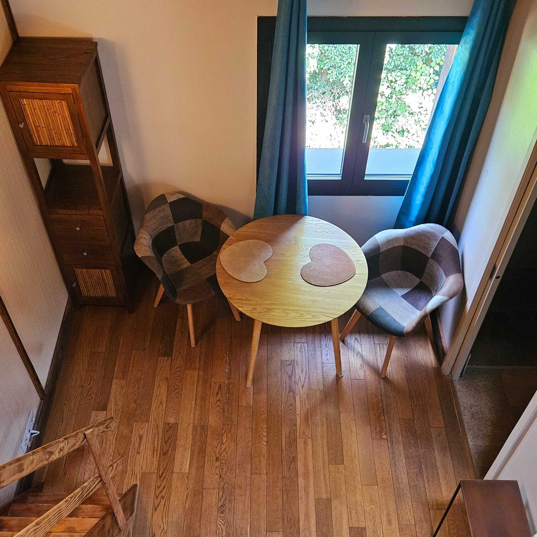 A corner room with a wooden floor, a round table with a cushion, two patterned chairs, a wooden cabinet, and a window with curtains.