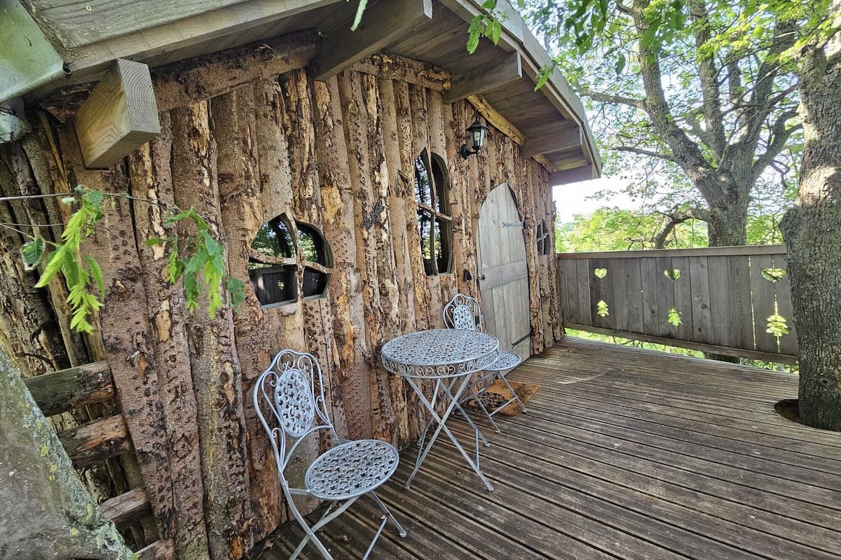 Een rustieke boomhut met een houten gevel, twee metalen stoelen en een kleine tafel op een houten terras.