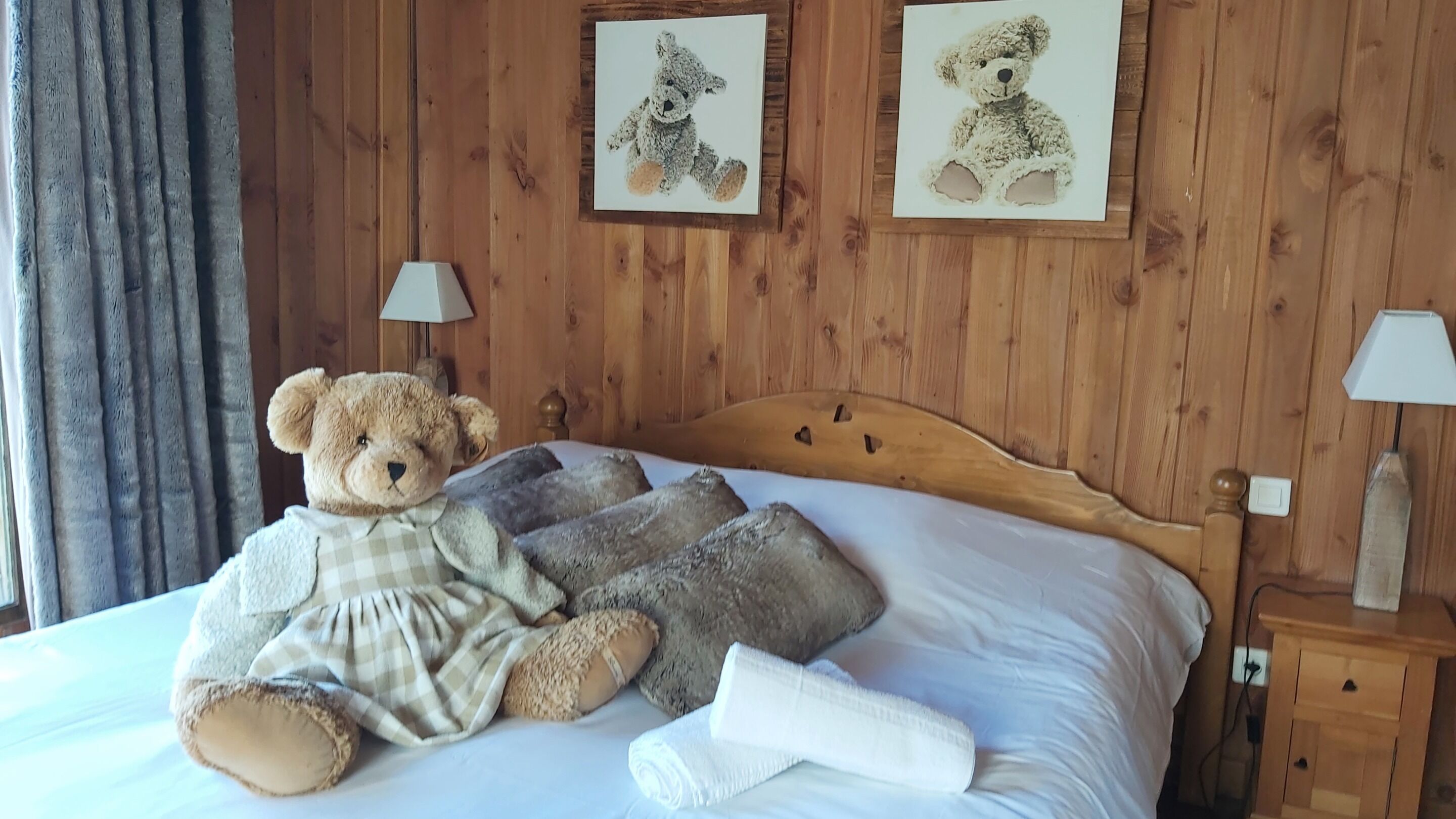 Een bed met een groot knuffelbeer, twee kussens, een deken en een opgerolde handdoek. Twee wandlampen en twee ingelijste afbeeldingen van teddyberen.